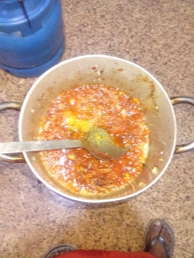 A top-down shot shows a pot of stew-like mixture, being stirred with a spoon. The location is identified as Aviation Quarters, Nigeria. A blue gas cylinder is visible on the top left, a person's foot clad in a sandal is partially seen on the bottom right, suggesting someone is cooking. The stew is in a metal pot with handles on the sides and a spoon. The surrounding floor is a mottled beige color.