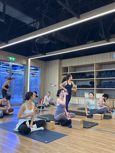 A yoga class is in session at a modern studio located in Nong Pa Khrang, Thailand. The room has light wood-paneled walls, a wooden floor, and a dark ceiling with industrial lighting. In the foreground, several women are seated on blue yoga mats, some in meditative poses. One woman in the center foreground wears a white tank top and black leggings, holding a phone. To her left, a woman in a purple tank top and leggings drinks from a water bottle. To her right, a woman in a teal top and grey leggings looks ahead, with two yoga blocks in front of her. In the background, a woman in a dark blue tank top and purple leggings stands holding two yoga blocks, facing a shelf unit stocked with rolled yoga mats and dark blue bolsters. Another woman in a white t-shirt and grey pants sits further back on the right. To the left, a woman in a black tank top and leggings walks past a glass door with blue neon lighting and a decorative frosted pattern. The overall atmosphere is calm and focused.