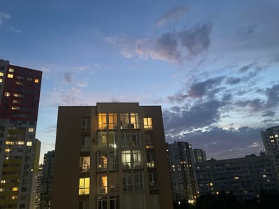 The image captures an evening view of apartment buildings in Brovary, Ukraine. Several multi-story residential buildings are visible, with lights on in many of the windows, indicating that residents are home. The sky is a twilight blue, with scattered clouds tinged with pink and purple hues, suggesting the sun has recently set. The buildings are varied in design and color, with some featuring balconies and air conditioning units. The overall scene depicts a typical urban residential area at dusk.