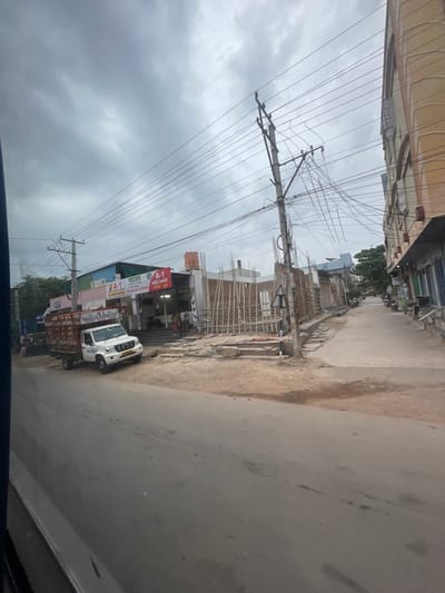 The image depicts a street scene in Bethamcherla, India, under a cloudy sky. A white pickup truck with a cargo bed is parked on the side of the road. The truck has "BROILER MARKETING" printed on its side. Adjacent to the truck is a building with a sign that reads "A-1". Further down the street, there are other buildings, some appearing to be under construction with wooden scaffolding. Electrical poles and numerous wires are visible overhead. The road surface is paved, with some dirt and debris on the sides. A few motorcycles are parked in the distance. The overall impression is of a commercial area with ongoing development.