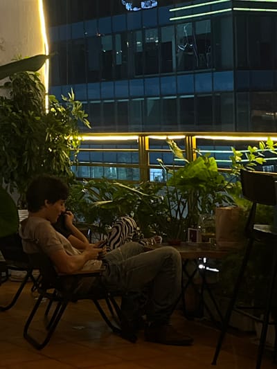 A person sits in a folding chair on a balcony at night, looking at their phone. They are wearing a light-colored t-shirt and blue jeans. To their right, a zebra-print fabric is visible. Behind them, lush green plants are illuminated by yellow lights. In the background, a modern building with blue glass windows stands tall. The balcony floor is made of wood. This scene is situated in Thủ Đức City, Vietnam.