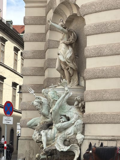 This media file captures a dynamic scene featuring classical sculptures and a partial view of a horse-drawn carriage. The primary subjects are two large marble sculptures, intricately detailed, depicting figures in motion. One sculpture on the left shows a reclining female figure with flowing drapery, seemingly reaching out. To the right, a more dramatic sculpture depicts multiple male figures engaged in a struggle, one holding a trident. The architectural setting features a curved, textured facade with large, vertical elements resembling pilasters or columns. Above and to the right, the head and tack of a dark brown horse are visible, suggesting the presence of a carriage.
The scene appears to be outdoors, possibly in a public square or courtyard in Vienna, Austria, given the architectural style and the hint of classical statuary. The lighting suggests it is daytime, though the precise time is not discernible. The sculptures evoke a sense of myth or historical narrative, with the intertwined figures conveying action and perhaps distress.
In the background, parts of buildings with windows are visible, along with a glimpse of a red-tiled roof, reinforcing the urban setting. A traffic sign with a red circle and a blue cross on a white background is prominently displayed, indicating a prohibition. Next to it, a white sign with black text reads "ENDE" (German for "END") and "11-22 ausgenommen 2 Fiaker." This translates to "11-22 hours exempt 2 Fiakers," likely referring to parking regulations related to horse-drawn carriages, a common sight in Vienna. The overall atmosphere is one of historical grandeur and everyday urban life coexisting.