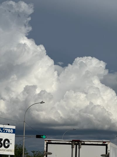 The image captures a dramatic sky filled with large, billowing cumulus clouds against a clear, grayish-blue backdrop. The clouds dominate the frame, with their bright white surfaces contrasting sharply with the darker, more somber clouds that stretch across the horizon. A portion of a white truck is visible on the right side of the frame, its metal exterior weathered and showing signs of age. Attached to a pole on the right is a traffic light displaying a green signal. Below the traffic light, a billboard-like sign is partially visible. The sign has a dark blue and white design with the number "7788" prominently displayed in white text against a blue background. To the right of this is a large "30" in black text. Text below this reads "МИНУТ" in black and "АВТО" in black. Further text below is illegible. The scene appears to be outdoors, possibly on a road, given the presence of the truck and traffic light. The lighting suggests it is daytime, with the sun likely casting harsh shadows, although these are not clearly visible due to the clouds. The overall mood is somewhat imposing, with the massive clouds and the starkness of the man-made elements creating a sense of scale and perhaps foreboding. The location is identified as Kokshetau, Kazakhstan.