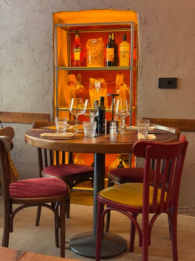 The image depicts the interior of a restaurant in Como, Italy. The setting is warm, with stucco walls and wooden accents. A lit recess in the wall displays bottles of alcohol (Aperol, limoncello) and packages of pasta. A wooden table is set with glassware. Red and brown chairs are arranged around the table. The overall impression is that of a cozy, Italian-style restaurant.
