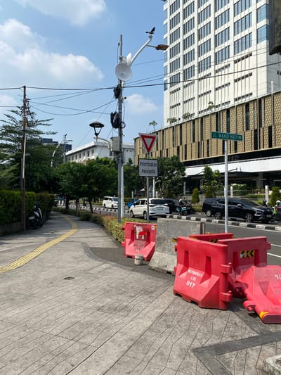 An outdoor, eye-level, medium shot captures a sidewalk and street scene in front of a modern, white building with many windows and an intricate, textured facade. The building appears to be located on Jl. Wahid Hasyim in Central Jakarta, Indonesia. The sidewalk, made of gray concrete with a patterned surface and a yellow tactile warning strip, curves to the left. On the right side of the image, red plastic barriers are stacked and placed along the edge of the street, indicating ongoing construction or road work. Concrete barriers are also visible.
A street sign reading "JL. WAHID HASYIM" is mounted on a pole. Another sign with a red triangle pointing down reads "Prioritaskan Pesepeda" (Prioritize Cyclists). A street lamp and various communication equipment are mounted on poles along the sidewalk and street.
The street itself is busy with traffic. A black car and a white SUV are visible, along with several motorcycles, one of which is parked on the sidewalk. Trees and dense green bushes line the edge of the sidewalk and the street. The sky above is bright blue with scattered white clouds. The overall impression is a sunny day in a developed urban environment.