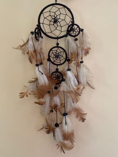 A dreamcatcher hangs on a light tan wall. The dreamcatcher is made of black thread, woven into a circular pattern, with smaller circular patterns within. Feathers of white and brown hang from the dreamcatcher, with black beads interspersed among them. The dreamcatcher is centrally located on the wall. There are no people or other objects visible in the image. The location is described as Kobuleti, Georgia, although this cannot be verified from the image alone.