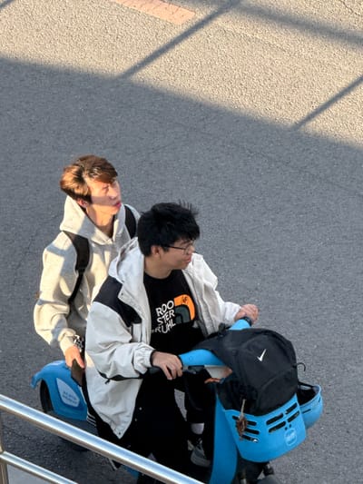 An overhead shot depicts two young individuals, possibly males, riding a blue tandem bicycle or scooter on a paved surface in Zhongtun, China. The scene is illuminated by strong, directional sunlight, casting long, distinct shadows across the ground, suggesting either early morning or late afternoon.

The person in the rear, with dark hair and glasses, wears a black t-shirt with white text that reads "ROOSTER RUHL" and a light-colored jacket with black accents. Their hands are on the handlebars of the blue vehicle. A black backpack with a white Nike swoosh logo rests on the back of the bike, alongside a blue helmet.

The person in front has lighter brown hair and also wears a light-colored jacket. Their face is partially illuminated by the sunlight.

Two blue helmets are visible, one attached to the back of the bike, and another to the front. The helmet in the rear clearly displays the brand name "AIRGO" in white lettering. The vehicle itself is predominantly blue, consistent with many shared bike services. The ground is a textured grey asphalt, with a lighter, sunlit section and darker shadowed areas. A metal railing is visible on the right side of the frame.