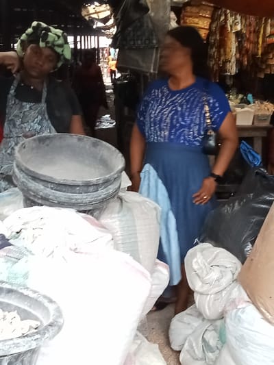 This image depicts a bustling, dimly lit indoor market scene in Owerri, Nigeria, characterized by crowded stalls and merchandise.

Two women are prominently featured in the mid-ground. On the left, a woman of apparent African descent wears a dark short-sleeved top, a patterned apron, and a green and black patterned headwrap. Her right hand is raised to the side of her head. She faces slightly right, appearing to look at the second woman. On the right, another woman of apparent African descent wears a blue short-sleeved top with a white abstract pattern, a plain blue skirt, and a small black shoulder bag. A light blue fabric is draped over her left hip, and she wears a gold ring on her left ring finger. She faces left, looking towards the first woman.

In the foreground and mid-ground, substantial quantities of goods are visible. A stack of four dark grey plastic basins, the top one containing a light-colored granular substance, occupies the left-center. Numerous large white and light-colored sacks, some with red or blue stripes, are piled around these basins and extending to the right, indicating bulk dry goods. Additional black plastic sacks are also visible on the right side of the frame. The background is blurred, showing a narrow market passage with a dimly visible figure further back. Hanging textiles or garments are discernible on the far right background. The market structure features visible roof supports and a potentially corrugated ceiling. The concrete floor shows signs of dampness in the central passage.