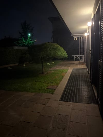 The image captures an outdoor nighttime scene, likely a pathway or corridor beside a building, in Lissone, Italy. The primary light source comes from artificial lamps, casting warm illumination on parts of the scene while other areas remain dark. The image appears to be rotated 90 degrees clockwise.

On the left side, a light grey or beige building wall features two square, modern outdoor wall lamps, casting light downwards. Below the lower lamp, a security camera or similar device is mounted. Above the wall, a dark architectural element with horizontal slatted patterns is visible, suggesting an overhang or part of the building's facade.

A dark tiled pathway, composed of large square pavers, runs along the right side of the image. Adjacent to this pathway, a long, dark metal grate extends parallel to the building, possibly for drainage or ventilation. To the left of the grate, a strip of green grass is visible. In this grassy area, a small, leafy tree with a slender trunk stands, and a few light-colored, indistinct objects are scattered on the grass.

Further into the background, another, darker wall made of large, textured blocks, resembling stone or brick, rises. Part of this wall appears to be covered by green vegetation or ivy. A metal structure, possibly a railing or support frame, is visible near the top of this wall. In the deeper background, a large, dark evergreen tree, likely a fir or spruce, is dimly lit, with a bright light source (possibly a distant streetlamp or reflecting light) nestled within its branches. The sky is dark, indicating a clear night.

No people are present, and no specific activity is taking place, creating a quiet, static atmosphere. There is no visible text in the image.