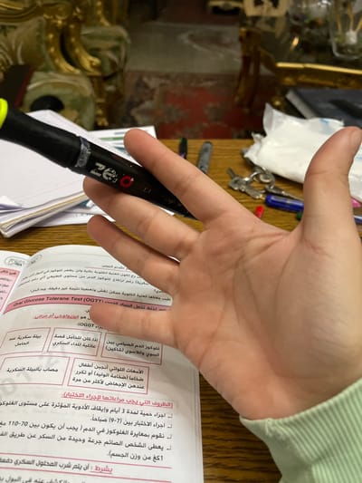 A light-skinned right human hand, wearing a light green long-sleeved garment at the wrist, holds a black highlighter pen with a yellow tip over an open textbook. The pen's body displays the word "zero" and a red "O" logo, with a white, worn band visible near the tip. The textbook, open on a light-colored page with Arabic text, discusses the "Oral Glucose Tolerant Test (OGTT)" and related medical conditions, with some text highlighted in pink. The scene is set indoors on a light brown wooden surface. In the blurred background, several objects are visible on the table, including a set of keys on a ring, a crumpled white plastic packet, and additional pens (one blue, one dark colored). Beyond the table, ornate gilded furniture and a red patterned rug are partially visible, suggesting a residential or study environment within the city of Sarouja Municipality, Syria.