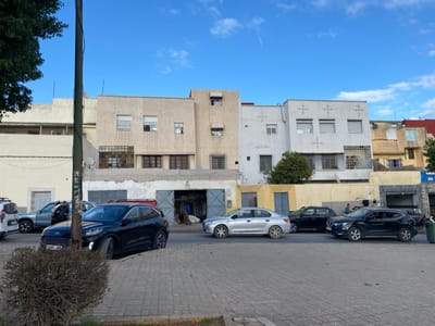 A street scene features several cars parked or moving on a paved road. Behind the vehicles are buildings, a mix of light-colored and beige structures with visible windows and varying architectural styles. One building appears to have a large opening, revealing interior goods. Above, a blue sky with scattered clouds completes the scene. A pole and some foliage are visible in the foreground.