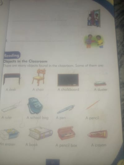 The image displays a page from what appears to be a children's book or educational material, featuring illustrations of classroom objects. At the top of the page, there are two small illustrations of children, one showing two children standing by an open door, and another of two children interacting. Below these, a heading reads "Reading" and "Objects in the Classroom". The rest of the page is dedicated to a grid of illustrated objects, each labeled with its name. These objects include: a desk, a chair, a chalkboard, a duster, a ruler, a school bag, a pen, a pencil, an eraser, a book, a pencil box, and a crayon. The page is white with some faint dotted lines suggesting a ruled or prepared surface. The overall aesthetic is that of an educational aid for young learners. There is no discernible connection to Jauro Yinu, Nigeria, or any urban environment in the provided image.