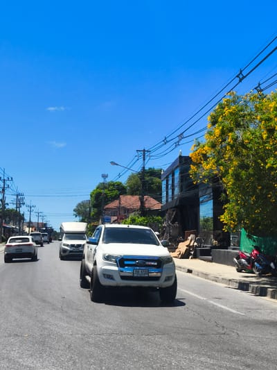 A low-angle, full-shot photograph captures a street scene in Baan Tai, Thailand, under a clear blue sky. The foreground is dominated by the front right quarter of a white Ford Ranger pickup truck, modified with oversized tires. The truck faces right and slightly forward, obscuring the road ahead. Further down the street, several other vehicles are visible, including a white SUV, a white van with a raised rear section, and a white pickup truck, all traveling in the same direction. The left side of the street features a row of buildings with utility poles and power lines running overhead. On the right side of the street, a modern, multi-story building with large, reflective windows stands out. Adjacent to this building is a vibrant green tarp covering stacked wood and other construction materials. A tree laden with bright yellow flowers is positioned in front of the building and tarp. Several motorcycles are parked to the right of the tarped materials. The road surface is asphalt with visible cracks and white lane markings. The overall impression is a sunny day in a commercial or semi-commercial area of Baan Tai.