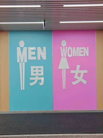 This image shows a pair of restroom signs for "MEN" and "WOMEN" on a wall. The "MEN" sign is on a blue background and features a white silhouette of a man and the Chinese character for "man" (男). The "WOMEN" sign is on a pink background and shows a white silhouette of a woman and the Chinese character for "woman" (女). Both signs are large and prominently displayed. Above the signs, there is a textured ceiling with linear lighting. Below the signs, there is a dark mat on the floor. The wall on the left and right of the colored sections is a neutral tan color. This scene is located in Fengtai District, China.
