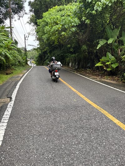 A person wearing a helmet is riding a scooter on a wet asphalt road in Karon, Thailand. The scooter has a black basket attached to the back, which contains some items. The road is bordered by lush green vegetation and trees on both sides. There are power lines visible overhead. The sky is overcast, suggesting it may have recently rained or is about to rain. The scene appears to be a quiet, rural or suburban area within Karon, Thailand.