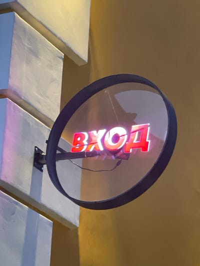 The image shows a close-up of a circular illuminated sign that reads "ВХОД" in Cyrillic letters, which translates to "ENTRANCE" in English. The sign is made of red neon or LED lights and is mounted on a dark frame. The sign is reflecting on a clear convex surface, possibly a mirror or a glass cover. The background consists of textured building facades, one in a light color with geometric angles, and another in a muted yellow hue. The lighting suggests it is nighttime or a dimly lit indoor environment. There are no people or animals visible in the image. The setting appears to be urban, with architectural elements typical of a city. The location is identified as Vologda, Russia.
