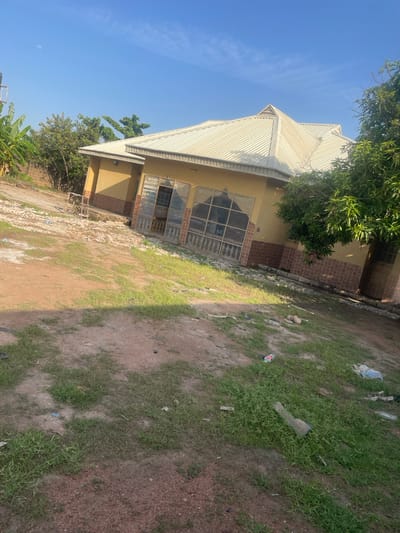 A residential building with a cream-colored corrugated metal roof is situated in an outdoor environment. The building has yellow walls with a decorative brown tile accent around the base of the structure. Multiple large windows with intricate metal grating are visible. The ground surrounding the building is a mix of dirt and patchy grass, with some scattered debris including plastic bottles and paper. Lush green trees, including banana plants and a large leafy tree, are present on the property. The sky is clear and blue with a visible moon. This scene is located in the city of Wauro Jabbe, Nigeria.