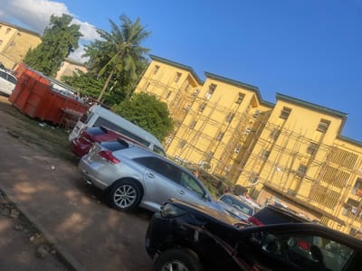 This image captures a bright, sunny day in Alagba, Nigeria, with a clear blue sky dominating the upper portion of the frame. The scene is set in an urban environment, likely a street or parking area in front of a large, yellow building. This building appears to be under construction or renovation, as evidenced by the extensive scaffolding surrounding it. Several cars are parked along the side of the road, including a silver sedan and a dark-colored car. A large, orange waste disposal container is also visible, with "LAGOS WASTE MA" printed on its side. Lush green trees, including a prominent palm tree, add a touch of nature to the urban landscape. The overall impression is one of an active, developing area on a beautiful day.