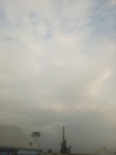 The image captures a cloudy sky above the city of Serti, Nigeria. In the foreground, a portion of a white, angled building is visible on the left, with some signage that is partially obscured. To the right of the building, a tall, thin smokestack or industrial structure stands against the hazy sky. Further to the right and slightly behind the smokestack, a solitary palm tree is silhouetted against the overcast backdrop. A faint power line can be seen stretching diagonally across the upper right portion of the frame. The overall atmosphere is subdued due to the heavy cloud cover.