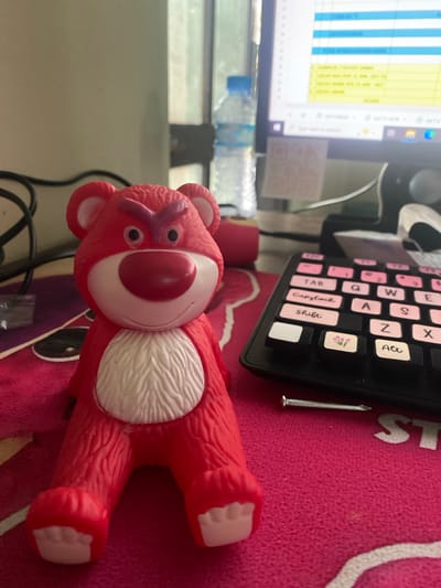 The image shows a close-up of a red stuffed bear toy placed on a pink textured surface. The bear has a large white oval muzzle with a red nose at the top. It has two small black eyes and pink x-shaped markings above them. White fur texture is visible on its chest. The bear's limbs are splayed out.
In the background, a portion of a computer monitor is visible, displaying a spreadsheet or table with blue and yellow columns. To the right of the monitor, a black keyboard with pink and white keys is seen. Some keys have handwritten labels, including "Capslock", "Shift", "Z", "X", "S", and "A". A small silver nail is placed near the keyboard. A plastic water bottle is also partially visible behind the monitor.
The overall scene appears to be an indoor workspace, possibly a desk, with the lighting suggesting it could be daytime. The setting is not clearly defined beyond the presence of a computer and keyboard. There are no people visible in the image, and the focus is on the toy and its immediate surroundings. The red color of the bear and the pink surface create a vibrant contrast. The image is a still photograph.