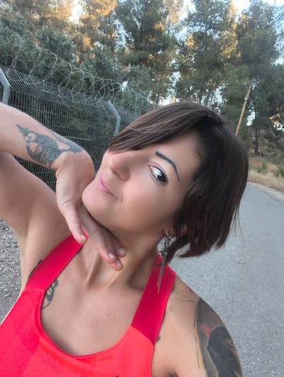 In a daylight outdoor scene, likely in the Lachish Regional Council, Israel, a woman with short, dark hair and tattoos on her arms and shoulder poses for a photo. She is wearing a bright red sleeveless top. Her left arm is raised, with her hand resting on her neck. She has a nose ring and is looking towards the camera with a slight smile. Behind her, a chain-link fence topped with barbed wire separates the area from a wooded hillside with trees. A paved road runs along the right side of the frame.