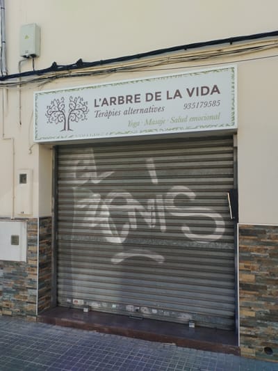 The image shows a storefront in Rubí, Spain. The business, called "L'ARBRE DE LA VIDA", offers alternative therapies, yoga, massage, and emotional health services. A sign with their name and phone number, 935179585, hangs above a closed metal shutter. Graffiti, indistinctly reading "eonis", is spray-painted on the shutter. The facade is a mix of plain wall and stone-like cladding. Utility boxes and wiring are visible above the storefront sign. A paved sidewalk is in front of the building.