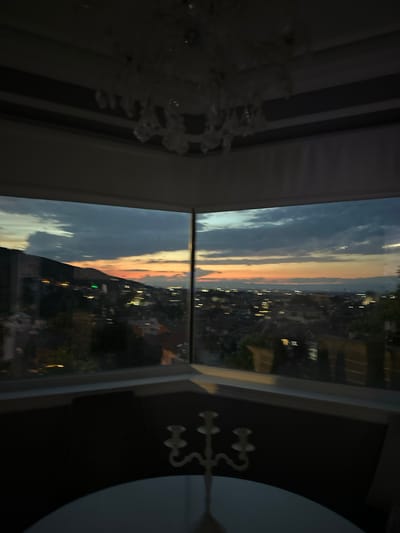 A wide-angle, low-light photograph captures a view of Asenovgrad, Bulgaria, from an elevated vantage point, possibly a residence or restaurant, through a large, corner window. The sky displays the colors of dusk with hues of deep blue, gray, and streaks of orange and yellow near the horizon, indicating a sunset. Scattered lights from the city below form a twinkling pattern against the darkening landscape. The foreground features the interior of the room, with a portion of a white table and a decorative candelabra visible. A chandelier with crystal accents hangs from the ceiling, partially obscuring the upper edge of the window frame. The overall impression is one of a serene evening cityscape viewed from a comfortable indoor setting.