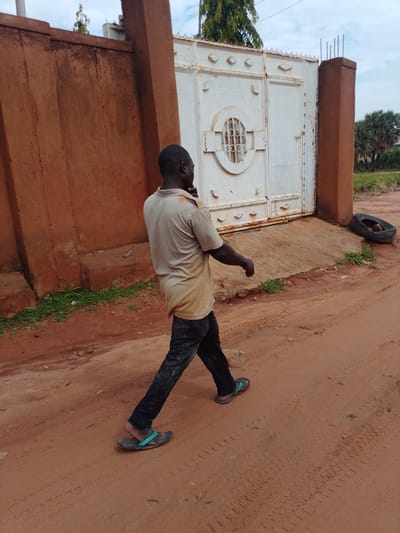 A man wearing a light-colored polo shirt and dark pants walks on a dirt road in front of a tall, reddish-brown wall and a white metal gate. The gate has decorative elements and a circular window with vertical bars. To the right of the gate, a discarded tire lies on the ground. The road surface shows tire tracks. Lush green vegetation is visible along the base of the wall and near the gate. The sky is overcast. The setting appears to be a residential area or compound.