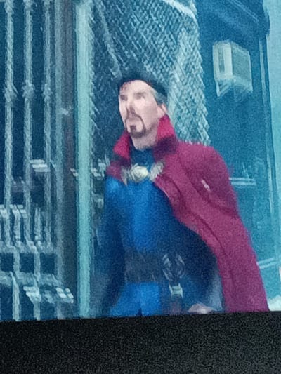 This is a medium shot, slightly pixelated image of a man dressed as Doctor Strange. He is wearing a blue tunic with a red cape draped over his shoulders. His head is slightly tilted down, and he appears to be looking to his left. He has a mustache and a goatee. He is standing in front of a blue and grey patterned background, which appears to be a building facade with some sort of grating or lattice work on the left side. There is a small, rectangular object mounted on the wall to the right of his head. The overall impression is a still from a movie or a promotional image. The context of Prague, Czechia, is not evident from the visual information in the image itself.