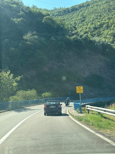The image shows a winding road on a sunny day with lush green trees on a hillside. A dark-colored car is driving on the road, and a motorcycle is ahead of it, also traveling on the road. A yellow sign is visible by the side of the road with the text "Most Sv. Stefan L=16 m" which translates to "Bridge of St. Stefan L=16 m". The road has a blue railing on the left side. The location appears to be near the town of Sveti Stefan in Montenegro.