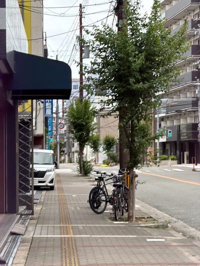 This media file captures an urban street scene in Osaka, Japan, from a slightly elevated perspective, likely during daytime under diffused lighting conditions.

The scene features a paved street on the right with an orange lane marking and white lines, bordered by a sidewalk equipped with yellow tactile paving. Lush green trees line the street, with several black bicycles chained to the trunk of one prominent tree.

To the left, multi-story buildings with varied architectural styles and grey and white facades rise. Overhead, numerous power lines crisscross the upper left portion of the frame. In the mid-ground, a white van is visible on the street.

The immediate foreground includes a dark, angular structure, possibly a utility box or part of a public fixture, alongside a metal railing with a diamond pattern.

There are no visible people or explicit activities, but the presence of bicycles and a van suggests a typical bustling city environment. Notable details include a green traffic light displaying a white arrow and a red circular road sign indicating a restriction (e.g., 'no entry' or 'forbidden').

Visible text on a blue sign in the mid-ground reads "大阪駅前橋" (Ōsaka-ekimae-bashi), which translates to "Osaka Station Front Bridge." Further text on the sign includes "マツモト" (Matsumoto) and the English word "KOBE."