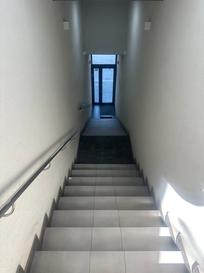 The image depicts an interior view of a modern, minimalist staircase and hallway in Ternopil, Ukraine. The scene is bathed in natural light streaming in from a large window or opening at the far end, suggesting it is daytime, likely with clear weather. The architecture is characterized by clean lines and neutral colors, primarily white and gray.
The primary subjects are the architectural elements themselves: a wide, gently sloping ramp or hallway leading to a staircase with concrete steps and gray tiled risers. A sleek, silver-colored metal handrail runs along the side of the ramp, curving upwards with the incline. The walls are painted white, and the ceiling is also white, with subtle shadows cast by the light.
In the distance, a large, dark-framed window or glass door reveals a glimpse of the exterior, with some light and faint shapes visible through it. There are a few small, white rectangular objects placed on the floor of the hallway, possibly construction materials or temporary markers.
The overall impression is one of quiet, uncluttered space, possibly a public building or a contemporary residential complex. There is no visible text in the image. The lighting creates a dramatic contrast between bright, sunlit areas and shadowed portions, highlighting the geometric shapes of the stairs and the space. The scene evokes a sense of calm and order.