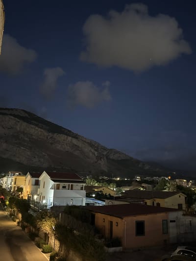 The image depicts a nighttime view of a residential area in Capaci, Italy, nestled at the foot of a large, rocky mountain. Several houses are visible, illuminated by artificial lights, with tiled roofs. A paved road runs along the left side of the frame, bordered by a stone wall and lush greenery. In the foreground, a small, peach-colored building with a dark window and some architectural details is prominent. The sky is dark blue, with scattered clouds, and a few faint stars are visible. The overall scene suggests a quiet, suburban evening in the Italian town.