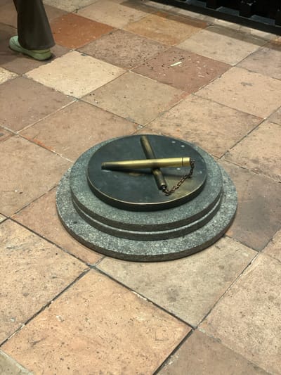 The image presents a high-angle view of a metallic, possibly brass or bronze, object resting on a tiered circular base, set into a tiled floor. The central object consists of a main vertical cylindrical piece with a short chain attached to its top. Two shorter, horizontal cylindrical bars extend perpendicularly from the base of the main cylinder, forming a cross shape. This assembly is placed on a dark, circular disk, which is then supported by two wider, concentric rings of light gray, speckled material resembling granite.

The entire structure is situated on a floor composed of square tiles laid out in a diamond pattern. These tiles vary in color from light brown to reddish-brown, showing some wear and texture. In the bottom left corner, the lower part of a person's leg, dressed in dark trousers and a light green shoe, is visible, indicating human presence.

The object appears to be a stationary fixture, possibly a historical or scientific instrument like a gnomon for a sundial, or a decorative marker. No specific activity is taking place beyond its static display. The lighting is even, suggesting an indoor setting or a well-lit covered area. Given the location context of Guang Zhou Shi, China, this could be part of a museum exhibit, a historical landmark, or a public space. There is no visible text in the image.