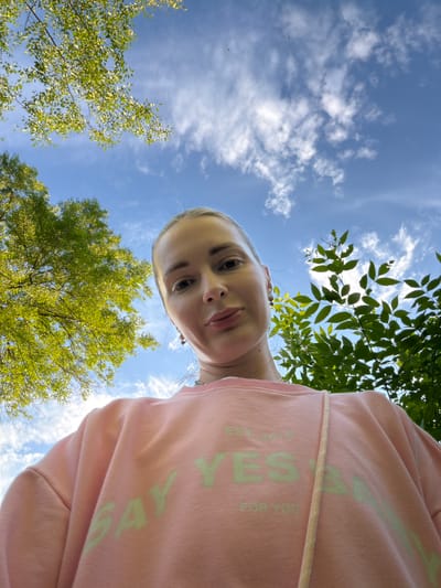 This is a low-angle, wide-angle photograph of a young woman looking up at the camera, taken outdoors during the daytime. The woman is positioned in the foreground, her face and upper chest visible. She has fair skin and blonde hair pulled back, and she is wearing a light pink hooded sweatshirt. The sweatshirt has a pastel green graphic on it that reads "SAY YES" in large letters, with "EST. 2019" and "FOR YOU" in smaller print below.
The background is dominated by a bright blue sky with scattered white, fluffy clouds. Green foliage from trees frames the top and sides of the image, suggesting a park or natural setting. Sunlight filters through the leaves, creating a dappled effect. The perspective implies the photo was taken from a low position, possibly lying on the ground or held above the head, looking upwards. The overall mood is bright and cheerful, with the woman's gaze directed towards the viewer. The presence of trees and open sky points to a warm weather season. The location context provided is Semiluki, Russia.
