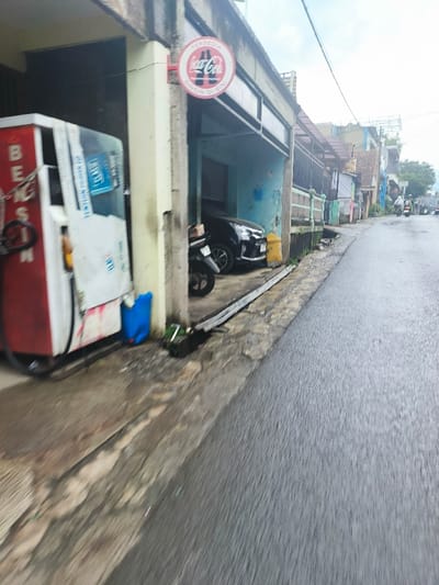 The image depicts a street scene in Cimenyan, Indonesia (latitude: -6.889, longitude: 107.651). A red and white gasoline pump sits on the edge of a paved street, partially obscured by a building. A black car is parked in front of the building, facing away from the viewer. A motorcycle is parked beside the car. A blue container sits near the pump. The street is wet and appears to have recently rained. Several buildings line the street on the right side of the image. A Coca-Cola sign hangs above the entrance to the building where the gasoline pump is located. The street is mostly empty, with no people visible. The buildings appear to be residential. The environment is overcast and cloudy.