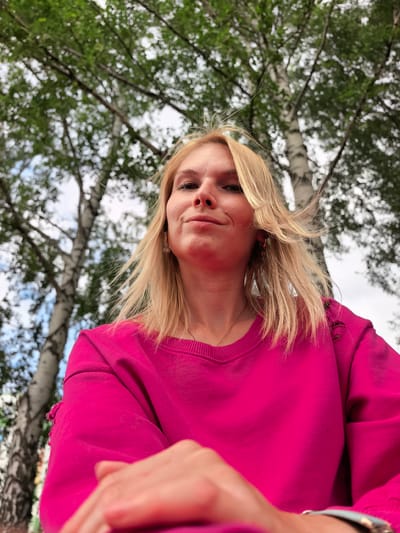 A low-angle, medium shot captures a young Caucasian woman with blonde hair and a smirking expression, wearing a bright pink sweatshirt, positioned outdoors amongst birch trees. Her hair is windswept, and she has a delicate gold necklace visible. The background consists of the white trunks of birch trees with green foliage and a cloudy sky. The woman's hands are visible in the foreground, one resting on her lap and the other slightly blurred in the lower right corner, adorned with a wristwatch. The image appears to be a selfie. The specific location in Sterlitamak, Russia cannot be determined from the visual information provided.