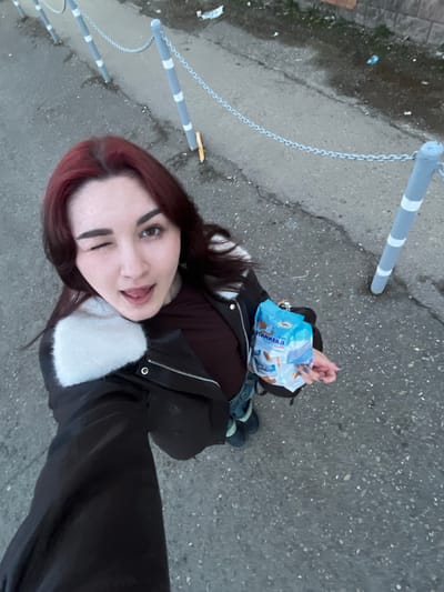 A young woman with dyed burgundy hair winks at the camera, standing on a gray concrete surface next to a row of gray and white striped bollards connected by chains. She is wearing a black leather jacket with a white fur collar, and holds a blue and white package of “Optimilk” wafer cookies. The background suggests an urban setting, with pavement, some vegetation, and a building visible in the distance. The photo has been taken from a high angle, looking down on the subject. Based on the packaging of the cookies, this scene is likely located in Russia. The specific city of Neftekamsk cannot be confirmed with this information alone, but the cookie brand and the general urban environment suggest this possibility.