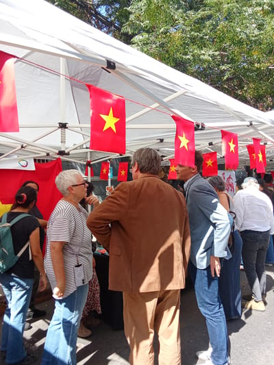 The image depicts a gathering of people under a white tent, likely an outdoor market or festival, in Providencia, Chile. Several individuals are engaged in conversations. One woman with short white hair and glasses, wearing a striped shirt and blue jeans, stands in conversation with a man in a brown suit. Another man in a blue blazer and jeans is also engaged in conversation nearby. The tent is decorated with small red flags, each featuring a yellow star, strung along its edges. The background reveals leafy trees, suggesting an urban park or tree-lined street setting. A sign with "BRUXELLES" can be seen in the background. The people appear to be browsing the market. The ground is asphalt.
