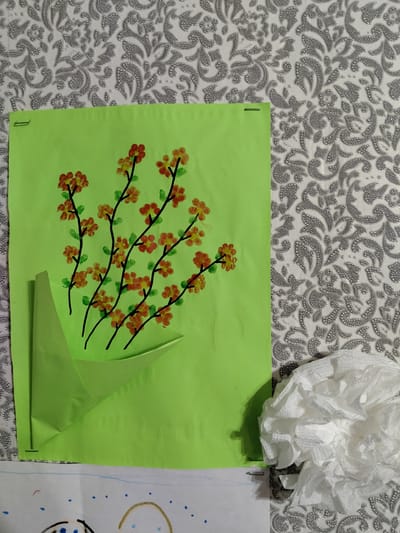 The image shows a piece of light green construction paper with a drawing of flowers. The flowers are made with orange and yellow thumbprints, with black stems and green leaves. The paper is attached to a grey and white patterned surface with staples. In the lower right corner, there is a crumpled white paper object, possibly a tissue. Below the green paper, another piece of white paper with drawings is visible. The location in the city of Luhansk, Ukraine is not discernible from the image.