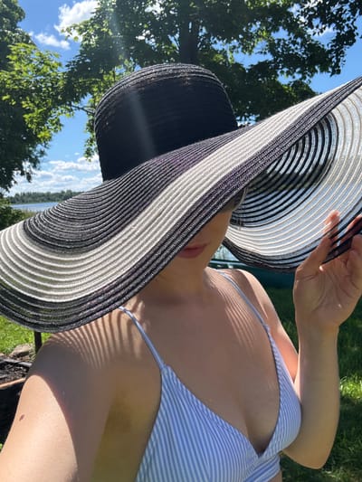 A woman wearing a wide-brimmed black and white striped hat and a blue and white striped bikini top poses for a selfie outdoors. The brim of her hat casts a shadow on her face, obscuring most of her features except for her lips and sunglasses. She holds the brim of the hat with her right hand. The background shows green grass, trees, and a body of water with a blue sky and white clouds. The scene appears to be a sunny day in a natural setting. The location is identified as Lendži, Latvia.