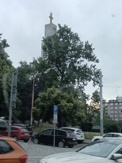 The image depicts a street scene in Prague, Czechia, under an overcast sky. A prominent church tower with a golden cross at its apex and clock faces is visible in the background. Several cars are parked along the street, including a red hatchback, a black minivan, and a white sedan. Trees with dense green foliage line the street, partially obscuring the view of a multi-story residential building in the distance. A blue and white parking sign is mounted on a pole. The street surface appears to be paved with cobblestones. There are no people visible in the image. The overall impression is that of a typical urban environment on a cloudy day.