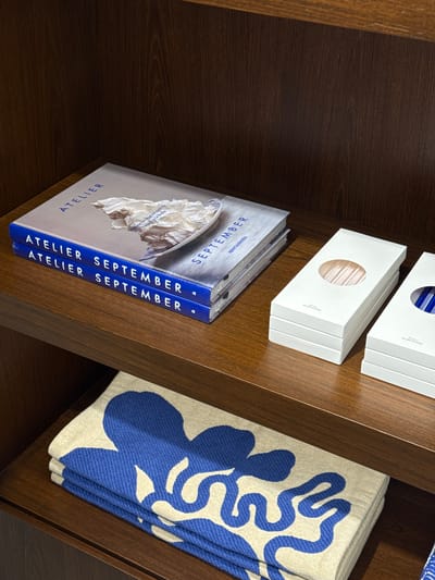 The image shows a close-up, angled view of a wooden shelf filled with various items. On the left side of the shelf, there are two white boxes stacked on top of each other. The top box is partially visible, and the bottom box has a circular cutout revealing a glimpse of what appears to be straws inside. Below these boxes, a stack of books or magazines is placed horizontally. The spines of these items are visible and clearly read "ATELIER SEPTEMBER" in bold blue lettering, repeated on each item. The cover of the topmost item in this stack features an image of white, crumpled material, possibly fabric or paper, against a light background.
To the right of the books, a folded textile is displayed, featuring a bold, abstract pattern of flowing blue shapes on a cream-colored background. The texture of the textile suggests it might be a blanket or a large scarf. The overall scene is well-lit, suggesting an indoor setting, possibly a retail display or a curated home environment. The wooden shelf has a rich, dark brown hue with a visible wood grain. The composition of the image, with its angled perspective and focus on the arrangement of objects, creates a sense of order and aesthetic appeal. No people are visible in the frame. The context provided states the location is Rome, Italy, but there are no specific visual cues in the image to confirm this.