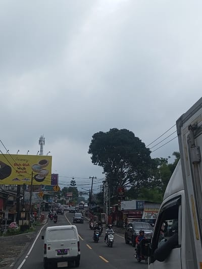 A road in Indonesia, perhaps near Puncak, is filled with cars and motorcycles on a cloudy day. A white van is visible on the right. A large billboard and cell tower rise up on the left. Shops line the road. A tall tree stands near the road. The sky is overcast.