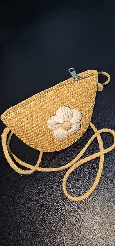 A straw-like woven bag with a rounded bottom and a zipper closure lies on a textured, dark surface. A pale yellow rope strap is attached to the bag and loops around it. The bag is decorated with a fabric flower made of shell-like pieces. The flower has a lighter center and petals that are a mix of cream and light tan. The zipper pull is silver. The overall impression is of a casual, summery accessory. The background appears to be a dark, possibly leather surface, with a visible texture. The lighting suggests an indoor setting, with the light source coming from above and slightly to the left, casting subtle shadows. There is no visible text. The image is likely a product shot or a personal photograph of the accessory.