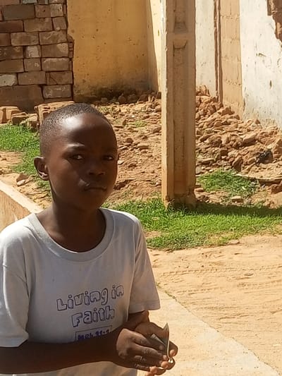 A young boy, likely of African descent, stands in front of a weathered, partially constructed building. He is wearing a light-colored t-shirt with blue lettering that reads "Living in Faith" and below it, "Heb 11:1". The background reveals a scene of rubble and dirt, suggesting ongoing construction or demolition. Sunlight indicates it is daytime, and the weather appears to be warm and dry. The boy's expression is neutral, and he is holding something small and metallic in his hands. The overall impression is one of resilience and everyday life in a developing or rural environment in Tempure, Nigeria.