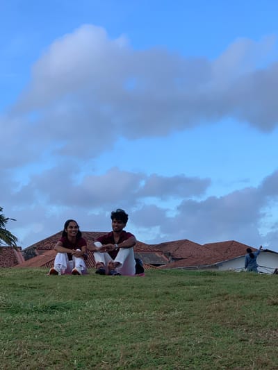 This is a photograph of a group of people on a grassy slope next to a building with a terracotta tiled roof. The sky is cloudy and appears to be either early morning or late afternoon, given the soft, diffused light. Two couples are visible in the foreground, sitting on a pink mat. They are both wearing maroon shirts and white pants. The couple on the right is looking towards the camera and smiling. Behind them, further up the slope, another person in a blue shirt is standing near the building. The overall mood of the image is relaxed and happy. The presence of palm fronds in the lower left corner suggests a tropical location, consistent with Galle, Sri Lanka.