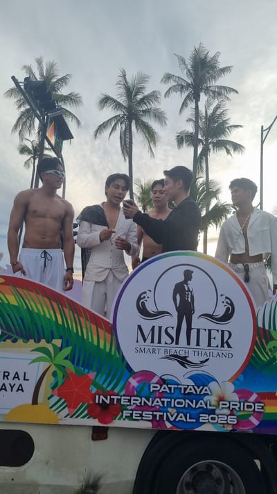 The image shows a group of young men participating in a pride festival parade. The setting appears to be outdoors, possibly on a street lined with palm trees, and the sky is overcast, suggesting it might be late afternoon or early morning. The participants are diverse in their attire, with some wearing white outfits that appear to be part of a costume, while others are shirtless or in casual wear. One individual in the foreground is shirtless, wearing white shorts and sunglasses. Another man, partially visible behind him, is also shirtless. Further back, a group of men are gathered around a large circular banner.
The banner is a prominent object in the image, featuring vibrant rainbow colors and various graphics. Text on the banner reads "MISTER SMART BEACH THAILAND" within a circular logo that depicts a silhouette of a muscular man. Below this, more text states "PATTAYA INTERNATIONAL PRIDE FESTIVAL 2025". The overall mood seems festive and celebratory, with the participants appearing engaged and in good spirits. The presence of palm trees and the text "Pattaya" strongly indicate the location as Pattaya City, Thailand. The event itself is clearly a pride festival, likely a parade or street celebration, given the presence of the banner and the gathering of people.
