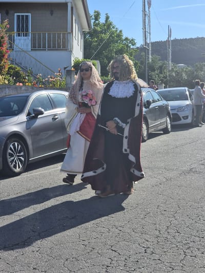 Two people are walking on a street next to parked cars. One individual is wearing a dark robe trimmed with white fur and a red cape, along with a curly wig and glasses. They are holding a silver object. The other person is dressed in a white gown with a red sash and a light veil, holding a bouquet of flowers. They are wearing sunglasses. In the background, there are houses, trees, and hills under a clear blue sky. Decorative poles are visible in the distance. The location appears to be in Barcelos, Portugal.
