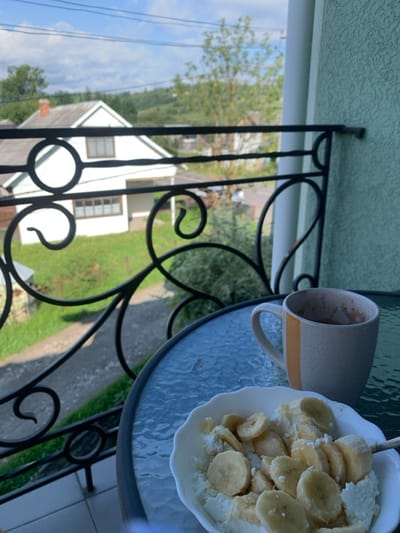 The image presents a daylight scene from a balcony in Lazeshchyna, Ukraine. In the immediate foreground, a round glass-top table holds a white, scalloped bowl containing cottage cheese topped with sliced bananas, and a two-toned grey and orange mug with a dark residue. A black, ornate wrought-iron railing with circular and scroll motifs extends across the mid-ground. Beyond the railing, a white two-story house with a reddish-brown roof and multiple windows is visible, situated on green grass with a dirt path leading away from it. The background features rolling green hills under a partially cloudy blue sky, with utility lines spanning horizontally. No individuals are present in the scene.